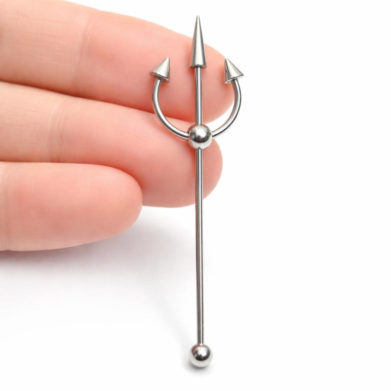May include: A silver-colored industrial barbell featuring a trident design. The barbell has a long, straight bar with a small ball at the bottom and a trident at the top, with a central spike and two curved spikes on either side.