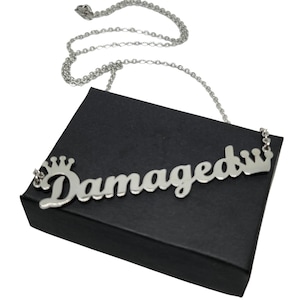 May include: Silver necklace featuring the word "Damaged" in cursive script with a crown on either side. The necklace is presented on a black box.