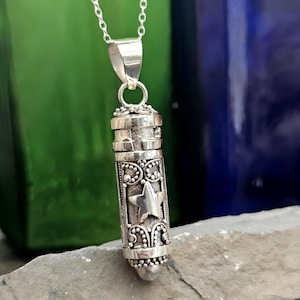 May include: A silver-colored pendant necklace with a cylindrical charm. The charm features a star design and ornate detailing. The necklace is suspended from a delicate silver chain, creating a classic and elegant look.