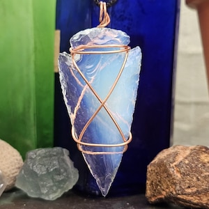 May include: A blue and white opal arrowhead pendant wrapped in copper wire. The pendant is hanging from a black cord.