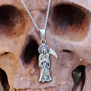 Pu&ograve; includere: Collana d'argento con un ciondolo dettagliato di una figura scheletrica che tiene una falce. Il ciondolo ha la forma di una persona che indossa una tunica.