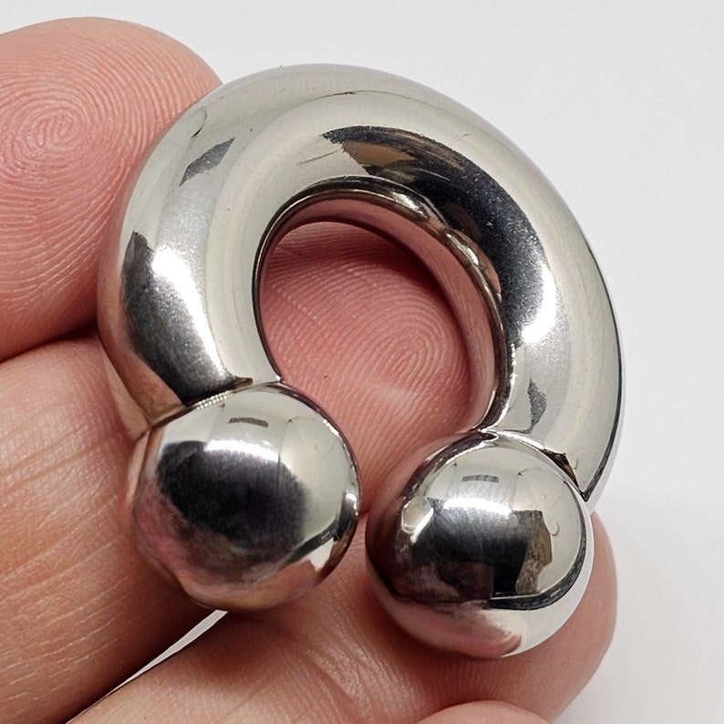 May include: A polished, silver-colored ear weight, also known as a saddle weight, with a curved design. The ends feature rounded, spherical accents. The jewelry is designed for stretched ear piercings.