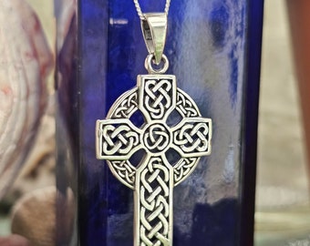 Puede incluir: Un colgante de cruz celta plateada con un intrincado diseño de nudos, suspendido de una delicada cadena. La cruz presenta un halo circular y patrones detallados, mostrando un estilo tradicional irlandés o escocés. El colgante se muestra sobre un fondo azul.