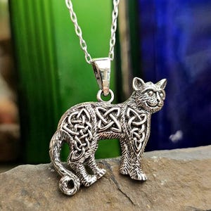 May include: A silver-colored pendant necklace featuring a stylized cat design. The cat has intricate Celtic knot patterns on its body and stands on a stone surface. The necklace chain is also silver.