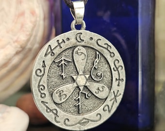 May include: A silver-colored pendant with a circular design. The pendant features a central star-shaped element with three stylized petals and various symbols around the edge. It hangs from a black cord.