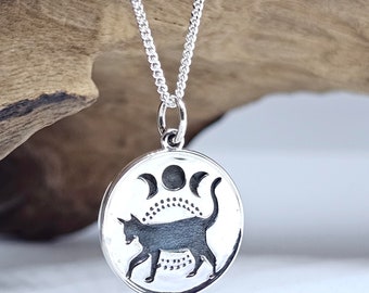 May include: A silver necklace with a round pendant featuring a black silhouette of a cat walking under a triple moon symbol.