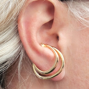 May include: Gold double hoop earrings worn in a person's ear. The earrings are made of two hoops, one slightly larger than the other, that are interlocked.