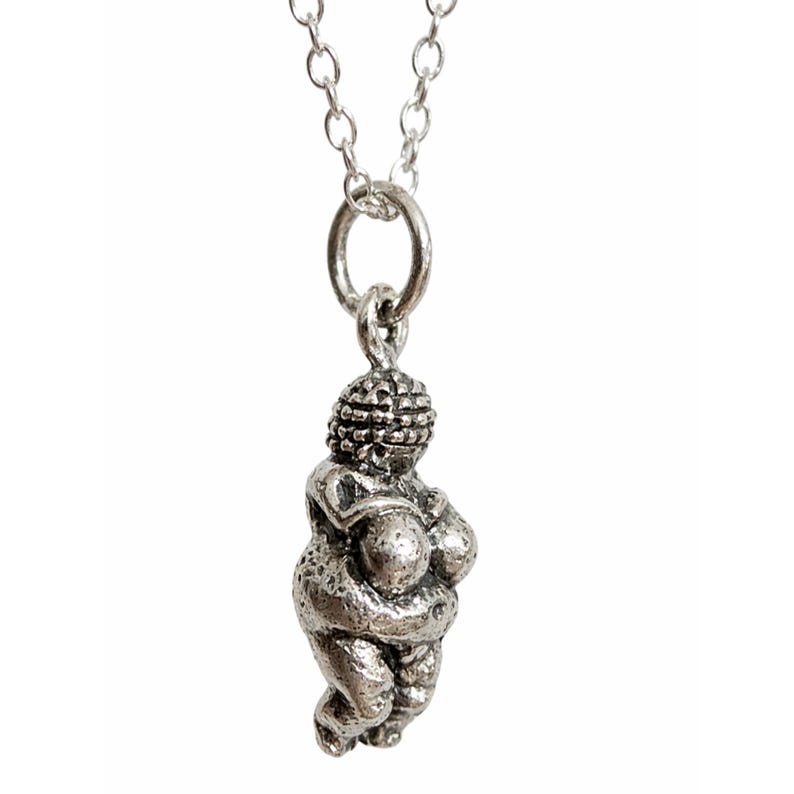 Puede incluir: Un collar con colgante plateado que presenta una peque&ntilde;a figura estilizada de una mujer. La figura tiene una forma redondeada con rasgos exagerados y est&aacute; suspendida de una delicada cadena.