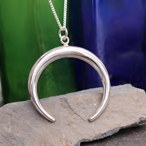 Può includere: Una collana con pendente a mezzaluna argentata. Il pendente ha una forma liscia e curva, sospesa a una delicata catena. La collana è esposta su una superficie di pietra grigia, con uno sfondo sfocato verde e blu.