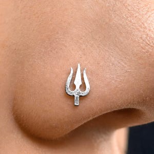 Pode incluir: Um pequeno piercing de nariz prateado com um design de tridente. A superfície metálica tem uma aparência texturizada e ligeiramente martelada. A forma de três pontas está centrada na pele, exibindo um acabamento metálico e um topo pontiagudo.