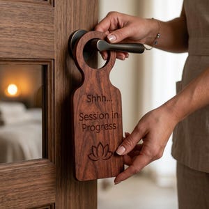 Pode incluir: Um cabide de porta de madeira com o texto "Shhh... Sessão em andamento" e um desenho de flor de lótus. O cabide de porta está pendurado numa porta de madeira castanha com uma maçaneta escura. A imagem sugere um ambiente de spa ou bem-estar.