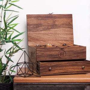 May include: A wooden box with a hinged lid and a pull-out drawer. The box is made of dark wood and has a natural finish. The lid is open and the inside of the box is empty. The box has a small metal latch on the front. The box has a drawer that is pulled out slightly. The drawer has a small metal handle. The box has the initials "BJ" engraved on the front.