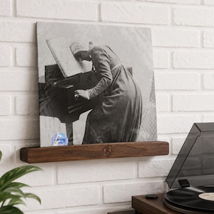 May include: A black and white print of a person playing a piano, displayed on a dark wood shelf. The shelf is mounted on a white brick wall. A record player is visible in the foreground.