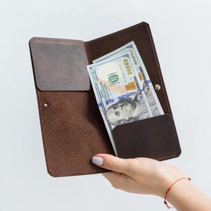 May include: A dark brown leather long wallet featuring a vertical orientation with visible stitching along the edges. The interior contains two dark brown leather pockets and holds several United States one hundred dollar bills.