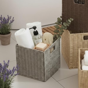 Może przedstawiać: Scena łazienkowa z kilkoma plecionymi koszami do przechowywania w odcieniach szarości, brązu i beżu. Kosze wypełnione są białymi ręcznikami, mydłem i akcesoriami kąpielowymi. Widać również wannę, świece i rośliny doniczkowe.