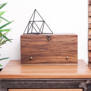 May include: A wooden box with a metal latch and two brass knobs sits on a wooden table. A black metal geometric pyramid sits on top of the box. A green plant is in the background.