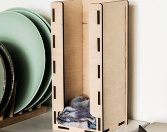 May include: A light-colored wooden dish towel holder with a rectangular shape and a slotted design. The holder contains a folded dish towel with blue, gray, and orange hues. Green plates are visible in the background.