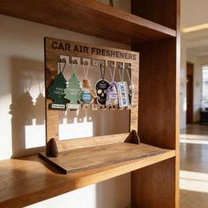 Könnte beinhalten: Holzständer mit der Aufschrift "CAR AIR FRESHENERS" und verschiedenen Auto-Lufterfrischern. Die Lufterfrischer umfassen Tannenzapfenformen, einen Totenkopf und ein Etikett mit der Aufschrift "NEW CAR SENT". Der Ständer steht auf einem Holzregal.