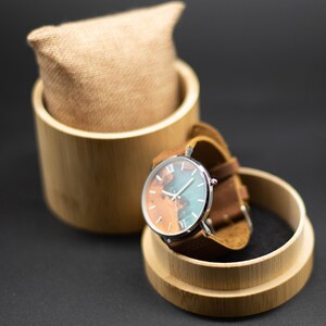 May include: A wristwatch with a brown leather band and a silver-colored case. The watch face features a unique design with shades of brown and teal. The watch is displayed in a bamboo box with a burlap cushion.