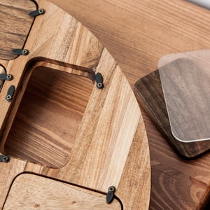 May include: A wooden tray with multiple compartments and a hinged lid. The tray is made of light brown wood with a natural grain pattern. The lid is secured with small black metal hinges and screws.