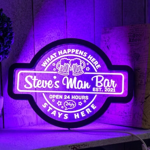 Peut inclure: Une enseigne au néon avec un cadre noir et du texte blanc qui dit "What Happens Here Stays Here". L'enseigne présente également deux chopes de bière, le texte "Steve's Man Bar Est. 2021", et "Open 24 Hours".