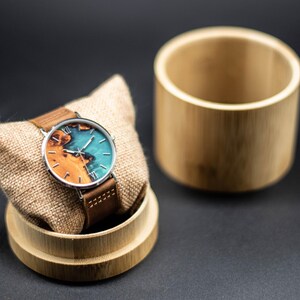 May include: A wristwatch with a brown leather band and a round face featuring a wood and turquoise design. The watch is displayed on a small, tan pillow inside a bamboo container, with a matching bamboo cylinder beside it.