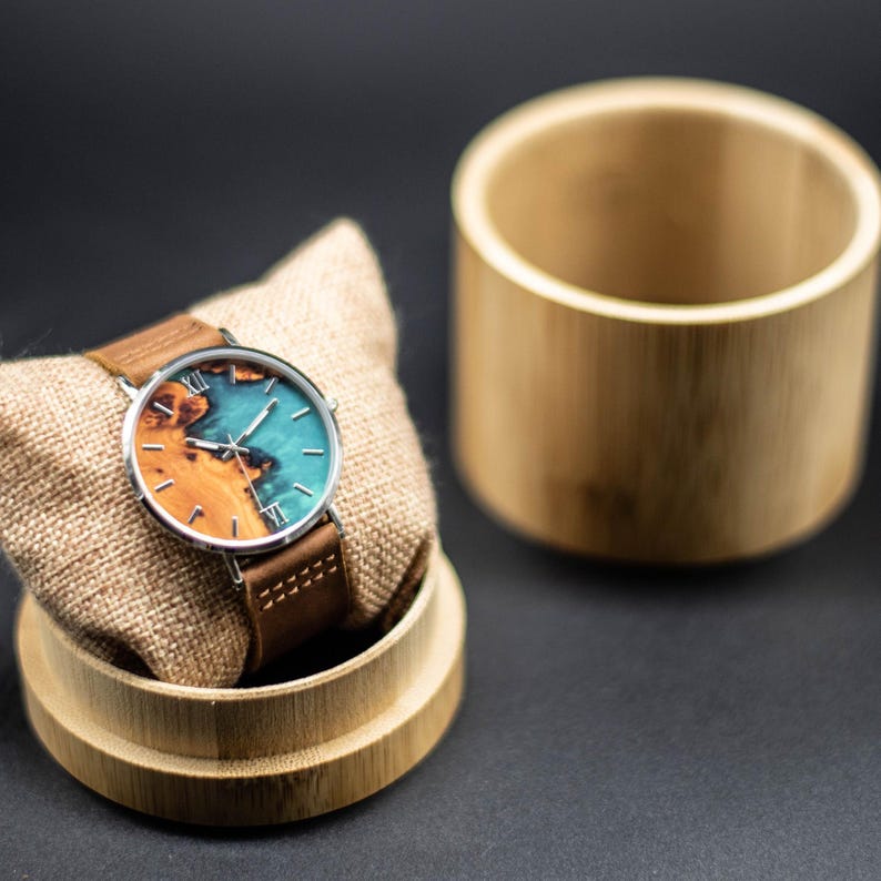 May include: A wristwatch with a brown leather band and a round face featuring a wood and turquoise design. The watch is displayed on a small, tan pillow inside a bamboo container, with a matching bamboo cylinder beside it.