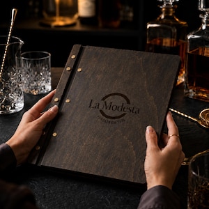 Puede incluir: Un menú de madera oscura, encuadernado con remaches de latón, se sostiene abierto. El menú presenta el texto "La Modesta" y "Fundada em 1918". El fondo incluye cristalería y botellas de licor.
