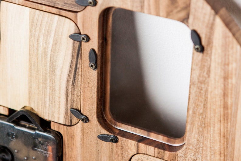 May include: Close-up of a wooden clock face with a metal clock mechanism. The clock face has a rectangular opening with a beveled edge. The clock is made of light brown wood with dark brown accents.
