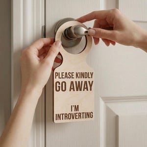 Peut inclure: Un accroche-porte en bois clair avec le texte "PLEASE KINDLY GO AWAY I'M INTROVERTING" en caractères gras. L'accroche-porte est tenu sur une porte blanche avec une poignée de porte argentée.