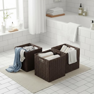 May include: Three dark brown woven storage baskets of varying sizes sit on a light beige rug in a bathroom. One basket contains folded clothes, while the others hold rolled white towels. The bathroom has white tile and a window.