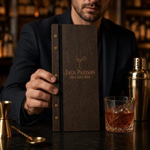 Op de afbeelding: Een donkerhouten menu met de tekst "Jack Parison For a Good Drink" en een cocktailglaspictogram. Het menu wordt voor een persoon gehouden. Een cocktailglas met ijs en een gouden cocktailshaker zijn ook zichtbaar.