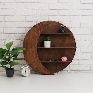 May include: A crescent moon-shaped wooden shelf with three shelves. The shelf is stained a dark brown and is mounted on a white brick wall. The shelf holds a small plant, candle, and other decorative items.