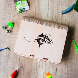Puede incluir: Una caja de madera con un diseño de pez, rodeada de aparejos de pesca. La caja tiene una tapa con bisagras y un gráfico de pez negro. Flotadores, anzuelos y señuelos de pesca de varios colores están dispersos sobre una superficie de madera.
