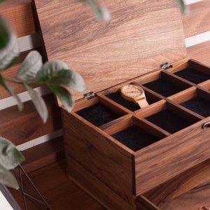 May include: A wooden jewelry box with a hinged lid and a black velvet interior. The box has multiple compartments for storing jewelry and a watch is placed in one of the compartments.