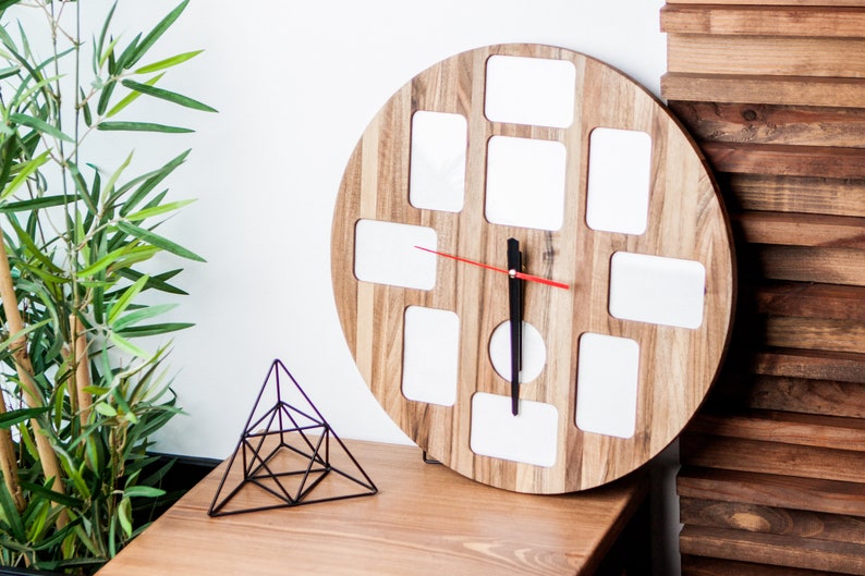 May include: A round wooden clock with 12 square picture frames. The clock has a black hour hand and a red minute hand. The clock is on a wooden table next to a black metal geometric sculpture.