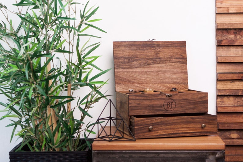 May include: A wooden box with a hinged lid and a pull-out drawer. The box is made of dark wood and has a natural finish. The lid is open and the inside of the box is empty. The box has a small metal latch on the front. The box has a drawer that is pulled out slightly. The drawer has a small metal handle. The box has the initials "BJ" engraved on the front.