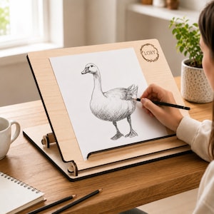 Op de afbeelding: Een houten tekenezel met een tekening van een gans. De ezel heeft het woord "LORY" in een cirkelvormig ontwerp. Een persoon tekent de gans met een potlood. De ezel staat op een houten tafel met potloden en een notitieblok.