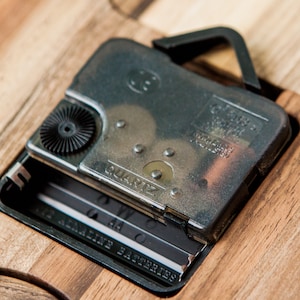 May include: Close-up of a silver clock mechanism with a black knob and the word "QUARTZ" printed on it. The mechanism is attached to a wooden surface with a black hook.
