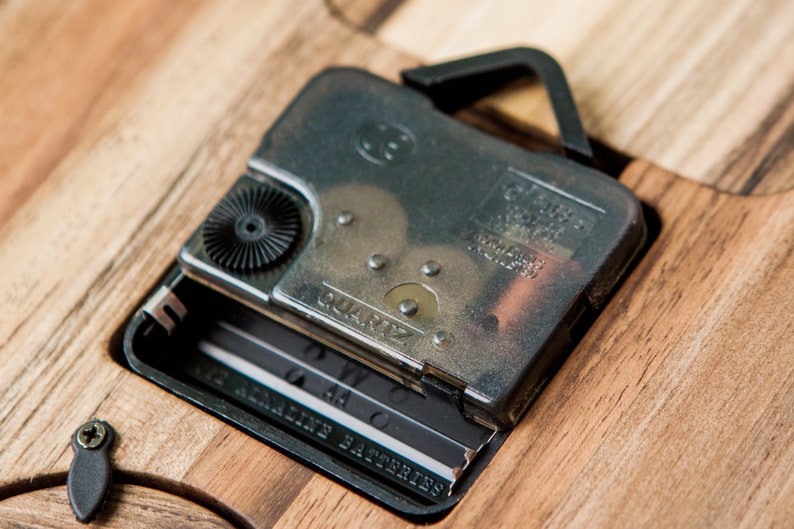May include: Close-up of a silver clock mechanism with a black knob and the word "QUARTZ" printed on it. The mechanism is attached to a wooden surface with a black hook.