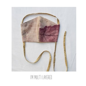 May include: A handmade face mask with a two-tone design. One side is a light beige, the other a deep burgundy with a leaf-like pattern. The mask has thin, mustard-colored ties. The text "I'M MULTI LAYERED" is printed at the bottom.