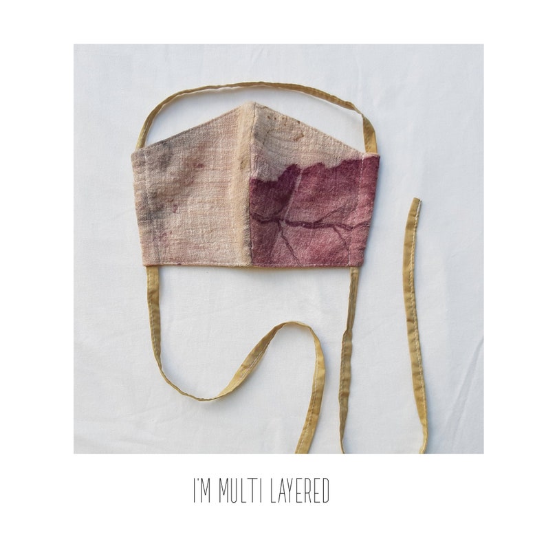 May include: A handmade face mask with a two-tone design. One side is a light beige, the other a deep burgundy with a leaf-like pattern. The mask has thin, mustard-colored ties. The text "I'M MULTI LAYERED" is printed at the bottom.