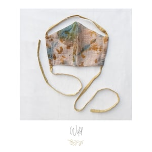 May include: A handmade face mask with a beige, blue, and brown pattern. The mask has long, gold-colored ties for securing around the head. The word "Wild" is written in cursive at the bottom of the image.