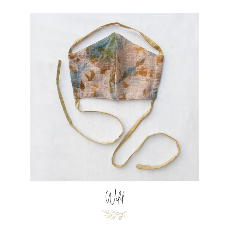 May include: A handmade face mask with a beige, blue, and brown pattern. The mask has long, gold-colored ties for securing around the head. The word "Wild" is written in cursive at the bottom of the image.