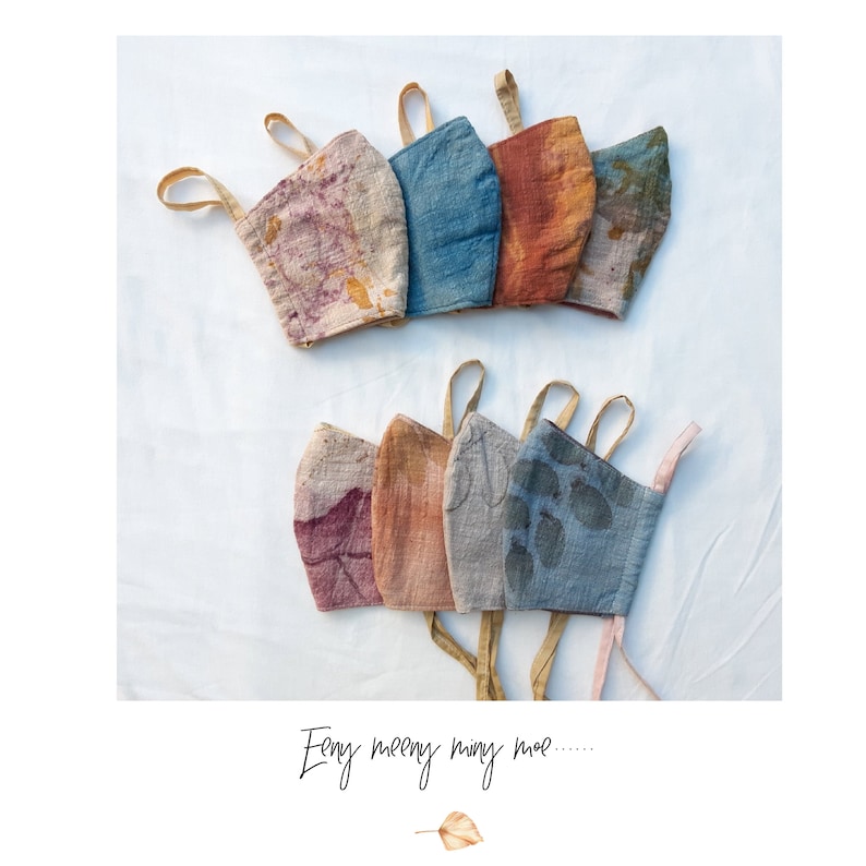 May include: A collection of handmade face masks in various colors, including purple, blue, orange, and gray. Each mask features a unique pattern and design, with fabric loops for securing around the ears. The text "Eeny meeny miny moe....." is written at the bottom.