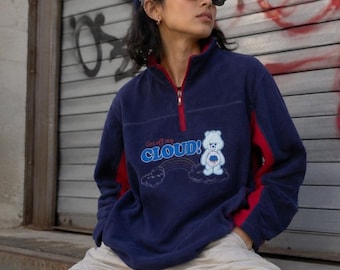 Könnte beinhalten: Ein marineblauer Fleece-Pullover mit Viertelreißverschluss, rotem Innenkragen und roten Seiteneinsätzen. Auf der Vorderseite befindet sich eine Grafik eines weißen Bären, eines Regenbogens und der Schriftzug Get off my CLOUD! in Blau und Rot.