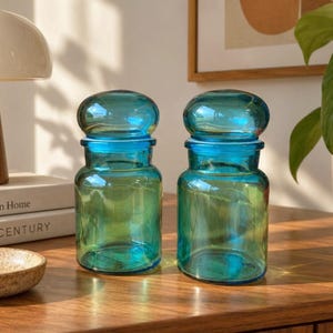 May include: Two matching glass jars with a vibrant teal blue hue. Each jar features a cylindrical body and a rounded, dome-shaped lid that sits securely on top. The glass is translucent, allowing light to pass through and create soft reflections.