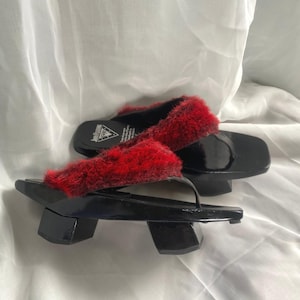 Puede incluir: Un par de sandalias geta negras con correas de piel roja. Las sandalias tienen un acabado brillante y un diseño tradicional japonés. La imagen muestra las sandalias sobre un fondo blanco.