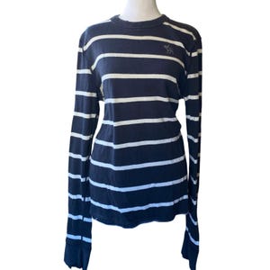 May include: A navy blue and white striped long-sleeved top. The top has horizontal white stripes on a navy blue base. A small logo is visible on the upper left chest. It has a crew neck and long sleeves.