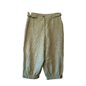 May include: High-waisted trousers in a textured olive green fabric featuring a single front button closure, integrated belt loops, and gathered cuffs at the bottom of each leg, each secured with a small dark button for a tailored finish.